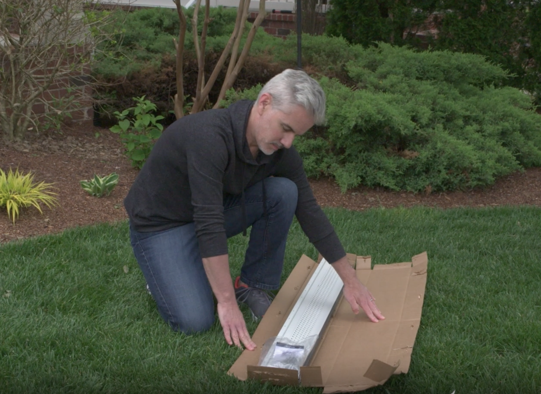 A man with gray hair kneels on grass, opening a large cardboard box containing a long white object—possibly featuring stainless steel micro-mesh—in a garden surrounded by shrubs, plants, and scattered pine needles.